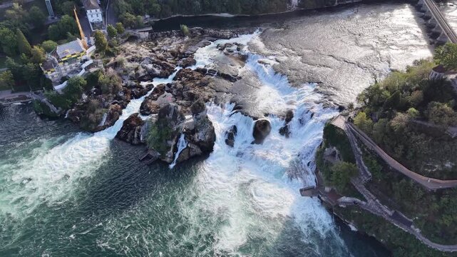 High aerial view of Rhine Falls in Switzerland, showing multiple cascades rushing around massive rock formations with cliffside viewing paths overlooking the turbulent water.