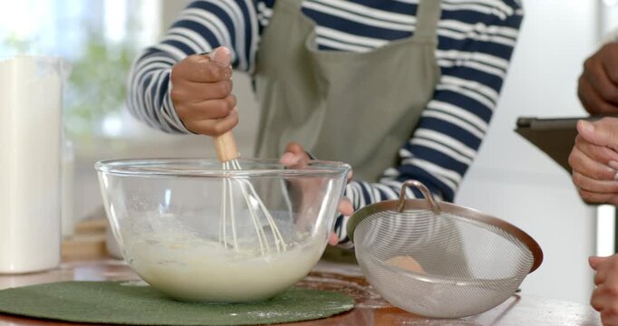 African American family in aprons guiding teen mixing batter for cake in kitchen using tablet whisk