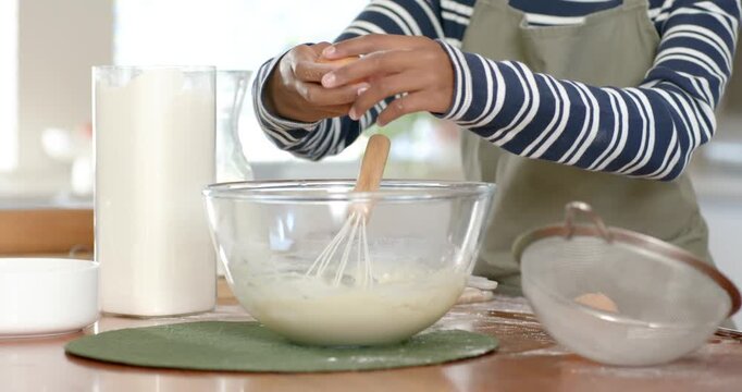 African American female cook holding egg, cracking into bowl at counter, whisking batter for baking