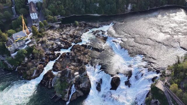 High aerial view of Rhine Falls in Switzerland, showing multiple cascades rushing around massive rock formations with cliffside viewing paths overlooking the turbulent water.