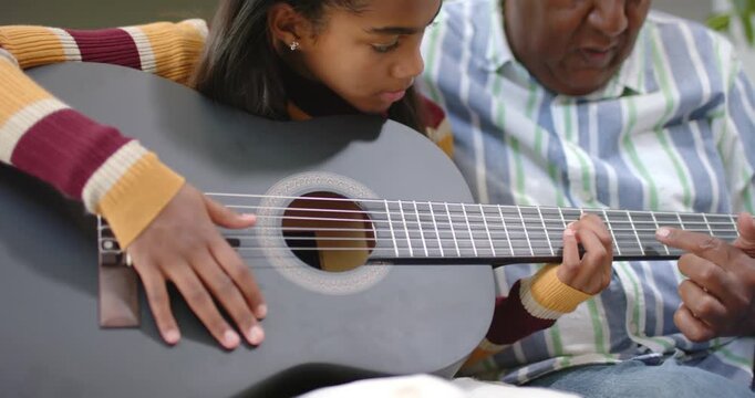 African American grandpa pointing to frets guiding girl learning chords on couch with black guitar