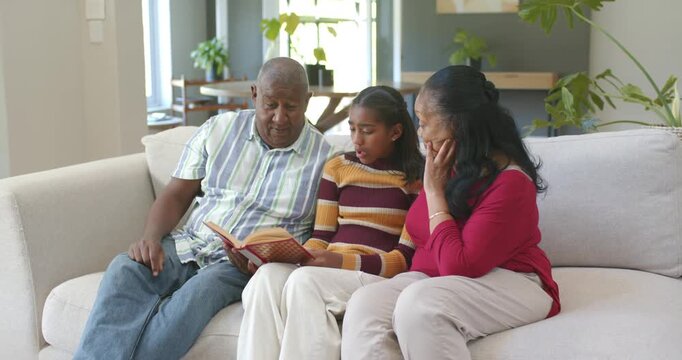 African family on sofa, teen girl leading shared reading while man following book, senior listening