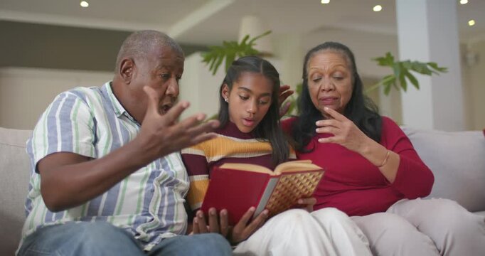 African American grandparents guiding granddaughter reading aloud after opening red book on sofa