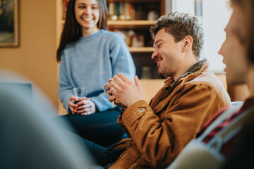 Three friends sit in a cozy living room, talking and laughing while holding drinks. Casual...