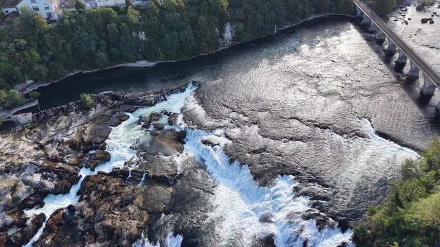 High aerial view of Rhine Falls in Switzerland, showing multiple cascades rushing around massive rock formations with cliffside viewing paths overlooking the turbulent water.