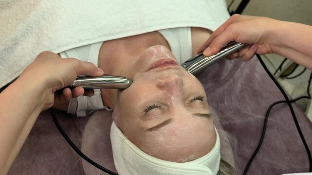 Beautician performs microcurrent skincare on a mature woman with dual facial probes, professional lifting, toning, rejuvenation and anti aging treatment.