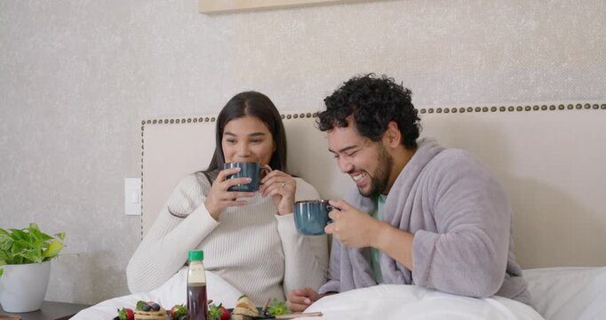 Wooden breakfast tray sparking African American couple laughing and sipping from blue mugs in bed