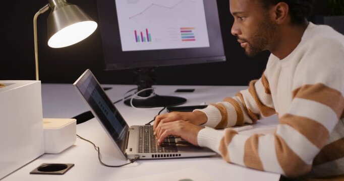 African American adult male at desk typing on laptop, comparing monitor charts and writing notes