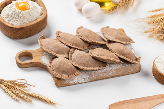 Raw uncooked mini chebureki meat pastries placed on wooden cutting board with flour and eggs on light table