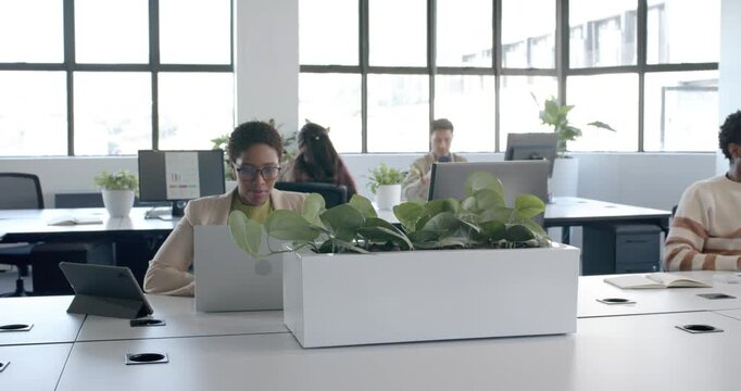 Diverse team in office camera panning revealing woman in blazer typing on laptop by planter working