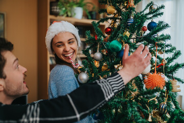 Naklejka premium A young couple decorates a Christmas tree while smiling and enjoying the holiday moment. The woman in a Santa hat laughs as they hang ornaments together by the decorated tree.