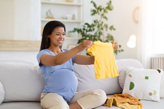 Happy pregnant Black woman sits on sofa at home, holding up a small baby shirt while organizing newborn clothes and preparing for the arrival of her child