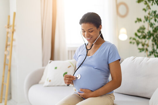 Happy pregnant Black woman sitting on sofa at home, listening to her baby bump with a stethoscope while smiling during a calm prenatal health and bonding moment