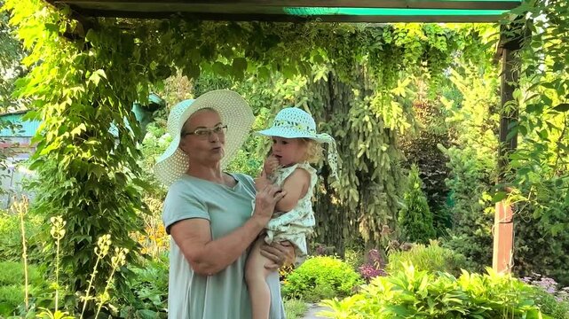 Senior woman carries her little granddaughter in a green garden, loving grandparent bond, family affection, childhood comfort and summer lifestyle.