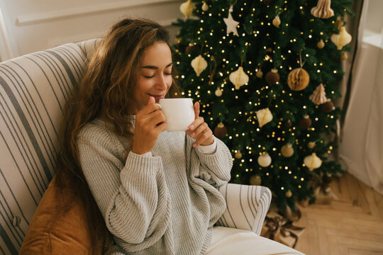 Young beautiful woman drinking tea sitting under decorated Christmas tree.