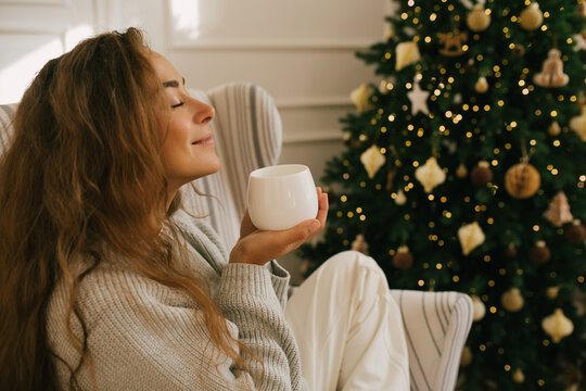 Young beautiful woman drinking tea sitting under decorated Christmas tree.