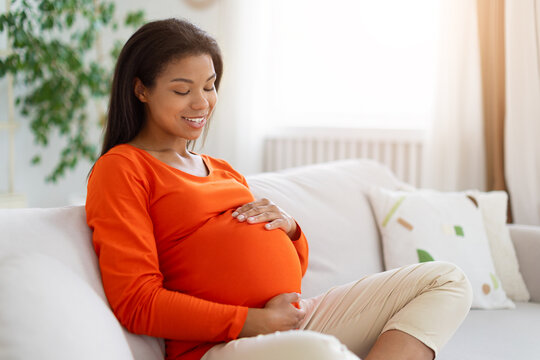 Smiling pregnant Black woman relaxing on sofa at home, gently holding her baby bump with both hands and enjoying a peaceful, comfortable moment during pregnancy