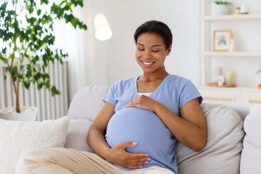Happy pregnant Black woman sitting on sofa at home, smiling while gently holding her baby bump with both hands and enjoying a calm moment in a bright modern living room