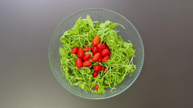 Hands dropping fresh red cherry tomatoes into glass bowl filled with green arugula leaves from above. Healthy salad preparation and organic Mediterranean cooking concept.