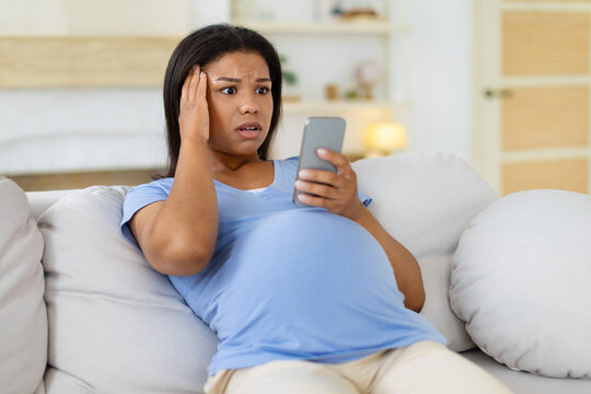Shocked pregnant Black woman reclines on a sofa at home, staring at her smartphone with a worried expression after receiving surprising or concerning digital news