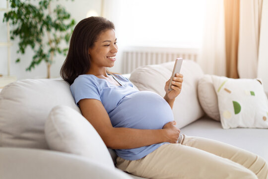 Happy pregnant Black woman relaxing on sofa at home, smiling while using smartphone and holding her baby bump during a comfortable everyday maternity moment