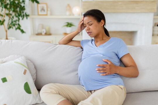 Pregnant black woman sitting on sofa at home with headache, holding forehead and belly, african american female showing fatigue and discomfort at home