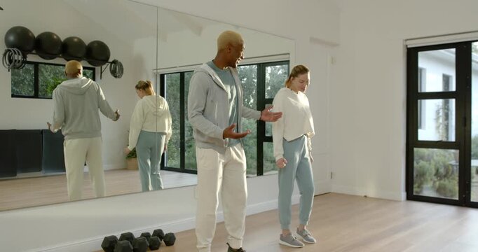 African American trainer gesturing, woman following, holding quad stretch for form at studio mirror