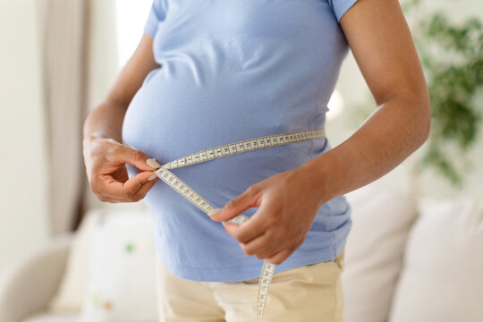 Close-up of pregnant Black woman measuring the circumference of her baby bump with tape measure at home, to track abdominal growth and pregnancy progress