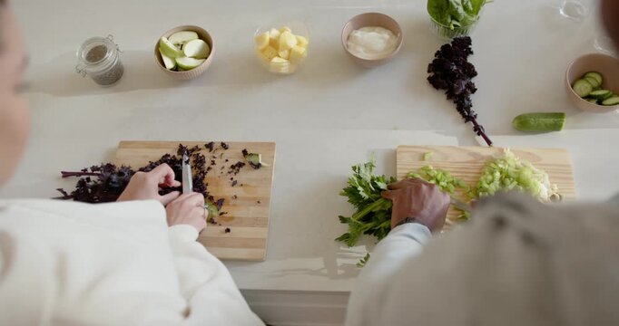 Couple chopping boards with knives at counter, ingredients set, woman guiding hand making salad