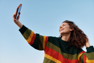 Fototapeta premium A happy young woman smiling and taking a selfie outdoors with her smartphone against a clear blue sky. Concept for social media, technology, youth.