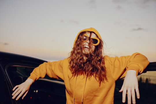 A young woman in a yellow hoodie and sunglasses leans happily against a car door at sunset. Concept for road trip, travel freedom, youth, and adventure.