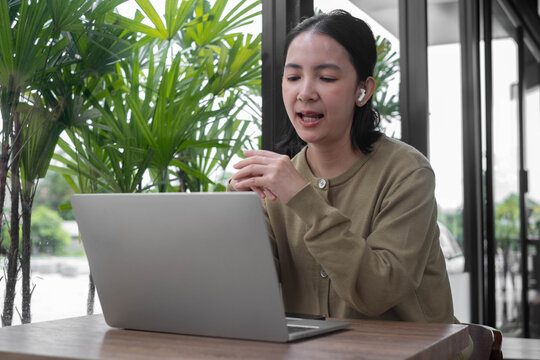 Asian woman using a laptop while talking through wireless earbuds in a bright modern workspace with tropical plants, representing remote work, online communication, productivity, and business.