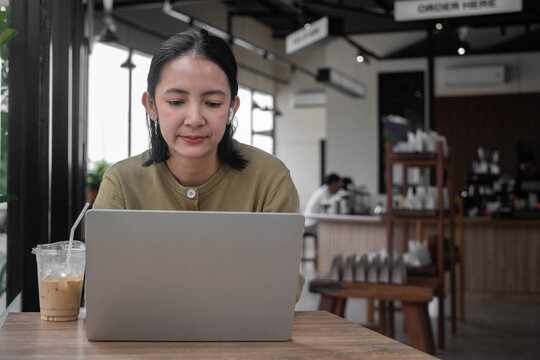 Asian woman concentrating on a laptop in a modern cafe, with wireless earbuds and iced coffee, representing remote work, focused productivity, digital lifestyle, online business, flexible employment.