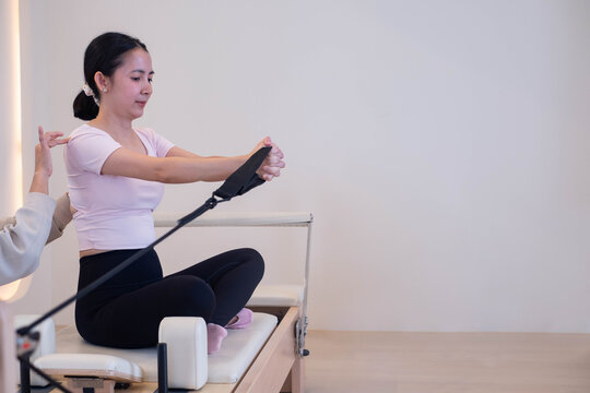 Woman practicing Pilates on a reformer machine in a bright fitness studio, stretching with a resistance strap, representing flexibility, strength, wellness, exercise, balance, and healthy living.