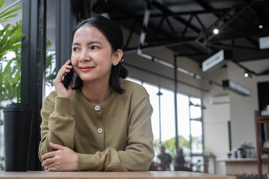Asian woman talking on smartphone in a cozy cafe, smiling while enjoying a coffee break indoors. Lifestyle scene showing communication, remote work, business, relaxation, and modern everyday life.
