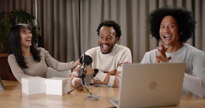 Diverse hosts laughing recording podcast center leaning mic left touching arm right checking laptop