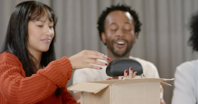 Friends watching woman in orange sweater opening cardboard box at table showing headset