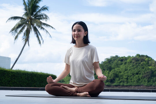 Asian woman practicing meditation outdoors in a tropical setting, surrounded by palm trees and lush nature, promoting mindfulness, relaxation, wellness, mental balance, self care, and healthy living.
