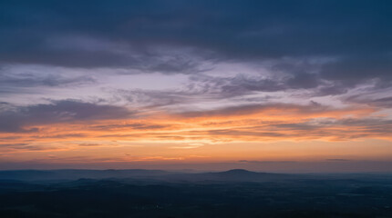 Fototapeta premium Panoramic Sunset Over Layered Hills and Distant Mountain Silhouettes with Vibrant Orange and Blue Sky, Representing Peaceful Landscape, Dusk, Nature Scenery and Golden Hour Atmosphere