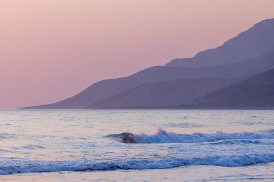 A tranquil seaside scene featuring gentle waves, a sandy shoreline, and a towering mountain. The setting sun paints the sky in warm, soothing hues. Kumluova Plaji, Patara, Turkey