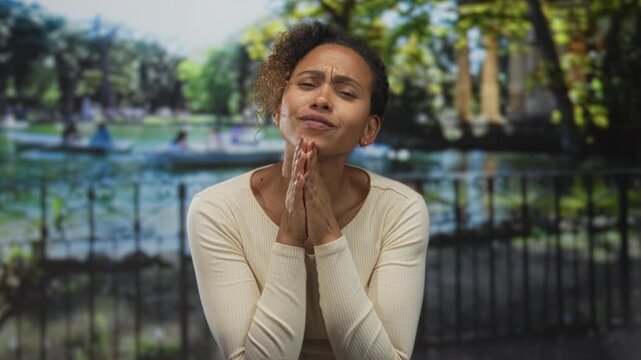Woman with hands clasped in prayer at a park railing beside a lake and paddleboats, eyes closed in pleading expression; hope resilience.