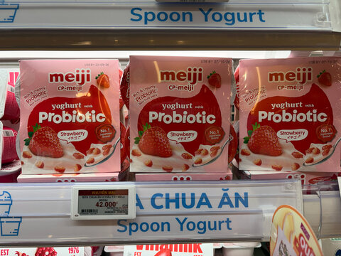 Three packs of Meiji probiotic yogurt arranged on a shelf. Each pack features a pink design with strawberry illustrations. The shelf is labeled 'Spoon Yogurt'.