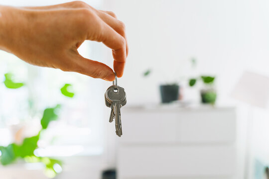 A hand holds new house keys against the blurred background of a bright modern apartment kitchen. Successful residential property purchase and exciting relocation commercial lifestyle concept.