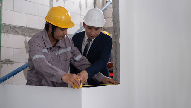 Construction workers and a site supervisor measuring an unfinished interior wall with a tape measure, showing teamwork, building inspection, safety, precision, quality control, and renovation work.