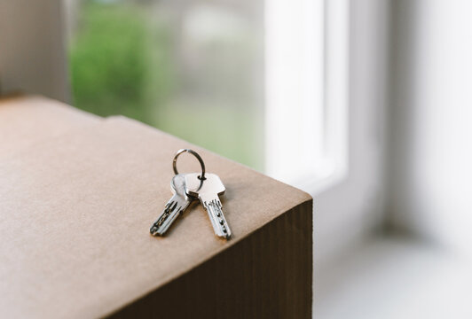 A shiny set of house keys lies abandoned on a cardboard moving box right next to a bright window. Authentic property ownership and exciting relocation transition lifestyle concept.
