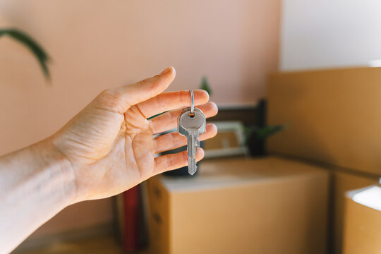 Hand holding keys with moving boxes indoors in new apartment. Relocation and residential property purchase commercial concept.