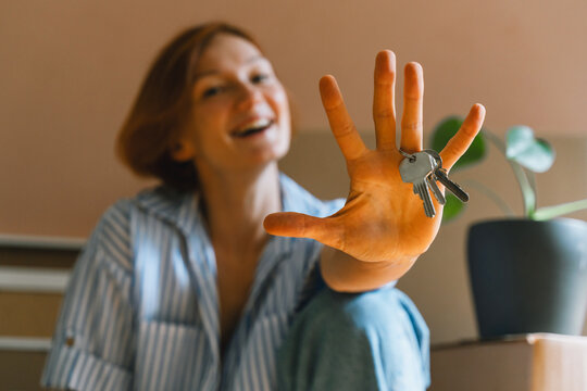 A happy Caucasian woman sits among cardboard boxes and playfully shows her new house keys to the camera. Authentic real estate ownership and successful relocation lifestyle concept.