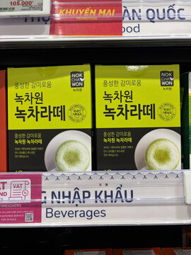 Green tea beverage packages are displayed on a shelf in a grocery store. The products are in bright yellow boxes featuring Korean text and a white cup with green tea. The setting is well-lit.