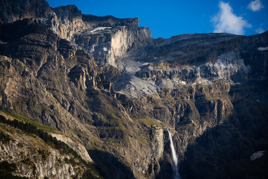 serene mountain landscape with cascading waterfall in alpine environment, aerial perspective showing rocky cliffs and sparse trees, brown and green tones dominate.