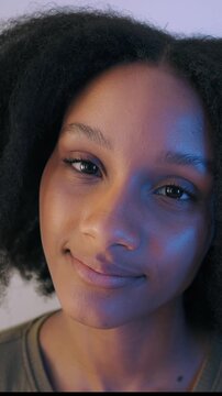 Black woman smiling at camera with curly hair and friendly expression, natural beauty portrait with visible skin texture and blue side lighting. Vertical video.